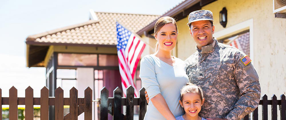 VA Home Loans provide a great opportunity for active duty or veterans to buy a home like this couple posing in front of their new house with an American flag.